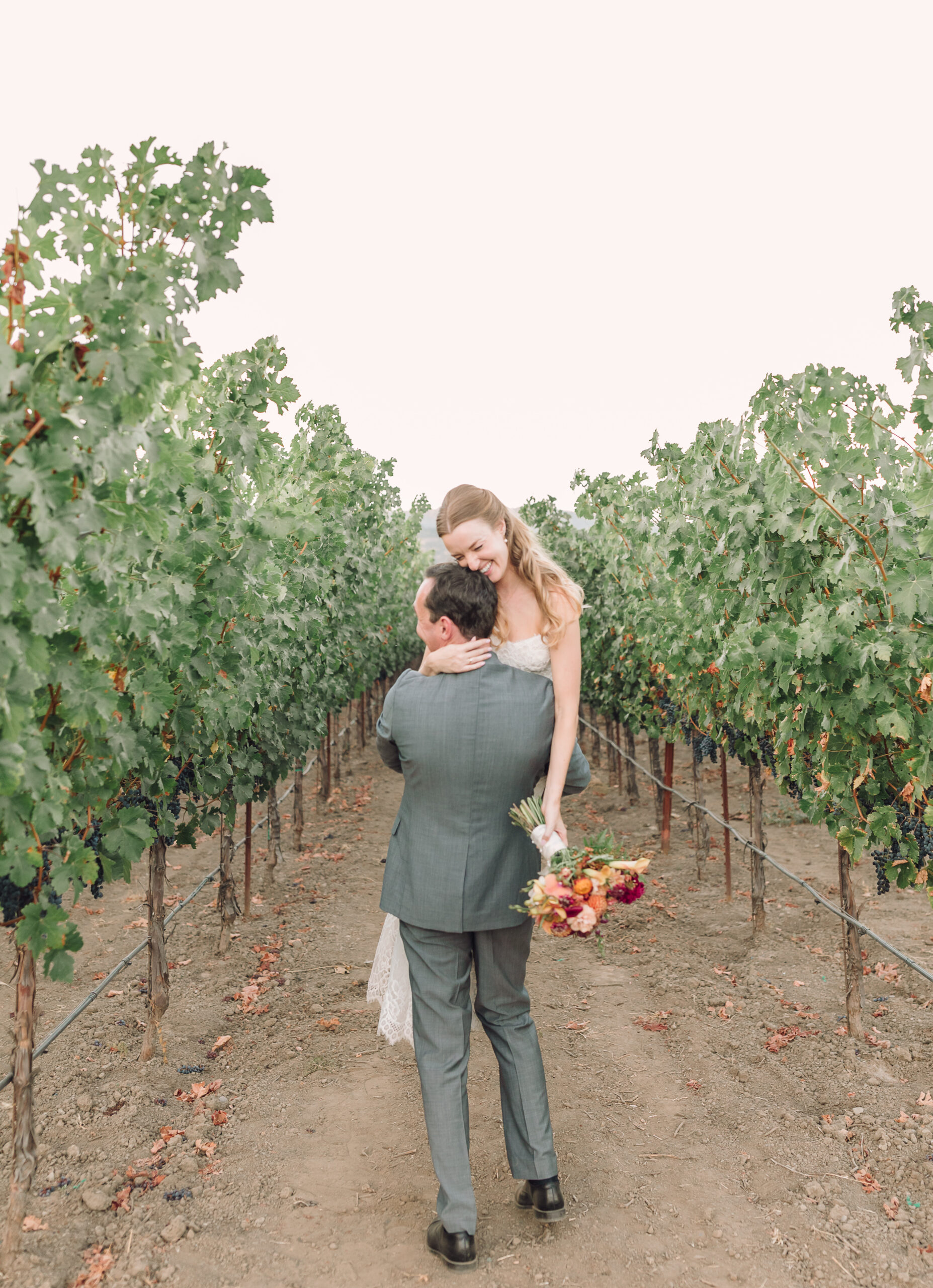 Bride and Groom at V. Sattui wedding in the vineyards at sunset. Groom playfully carries bride through the vineyard row.
